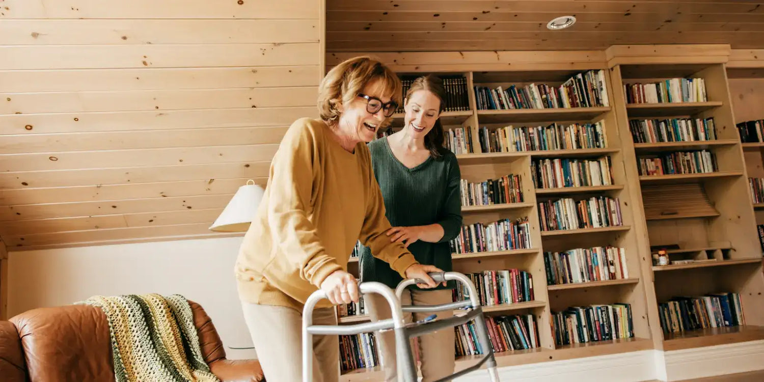 An older woman using a walker indoors, assisted by a younger woman in a cozy room with wood-paneled walls and bookshelves.