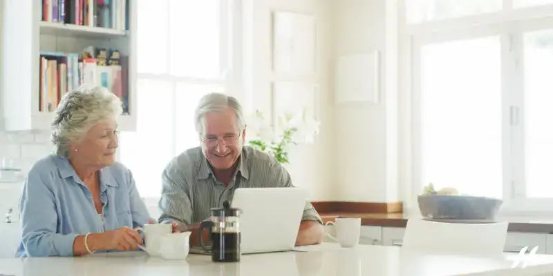 An elderly couple sitting at a kitchen table with a laptop, mugs, and a coffee press in a bright, modern kitchen.